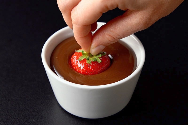 hand dipping a strawberry into melted chocolate using the stem