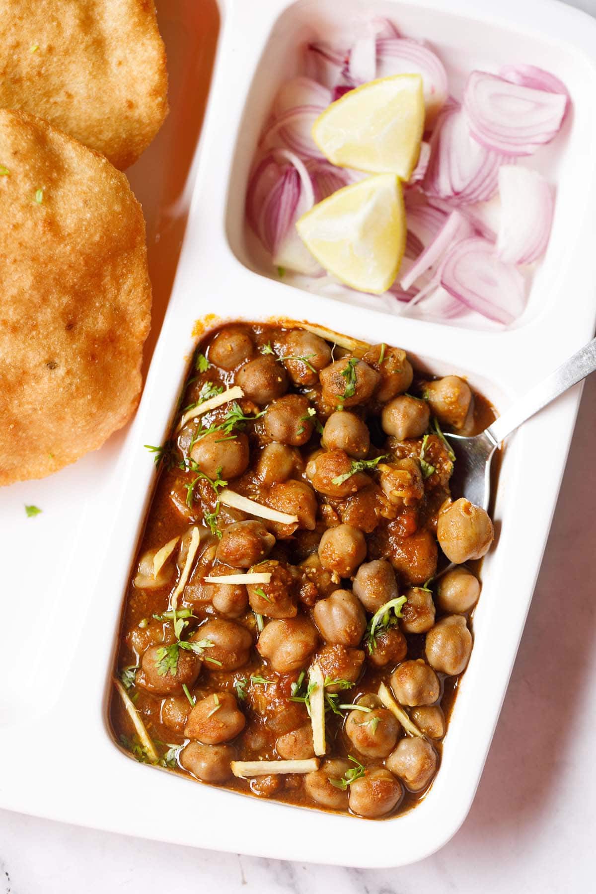 white plate with sections filled with chole masala, sliced onions, fresh lime wedges and bhatura on a marble table-top