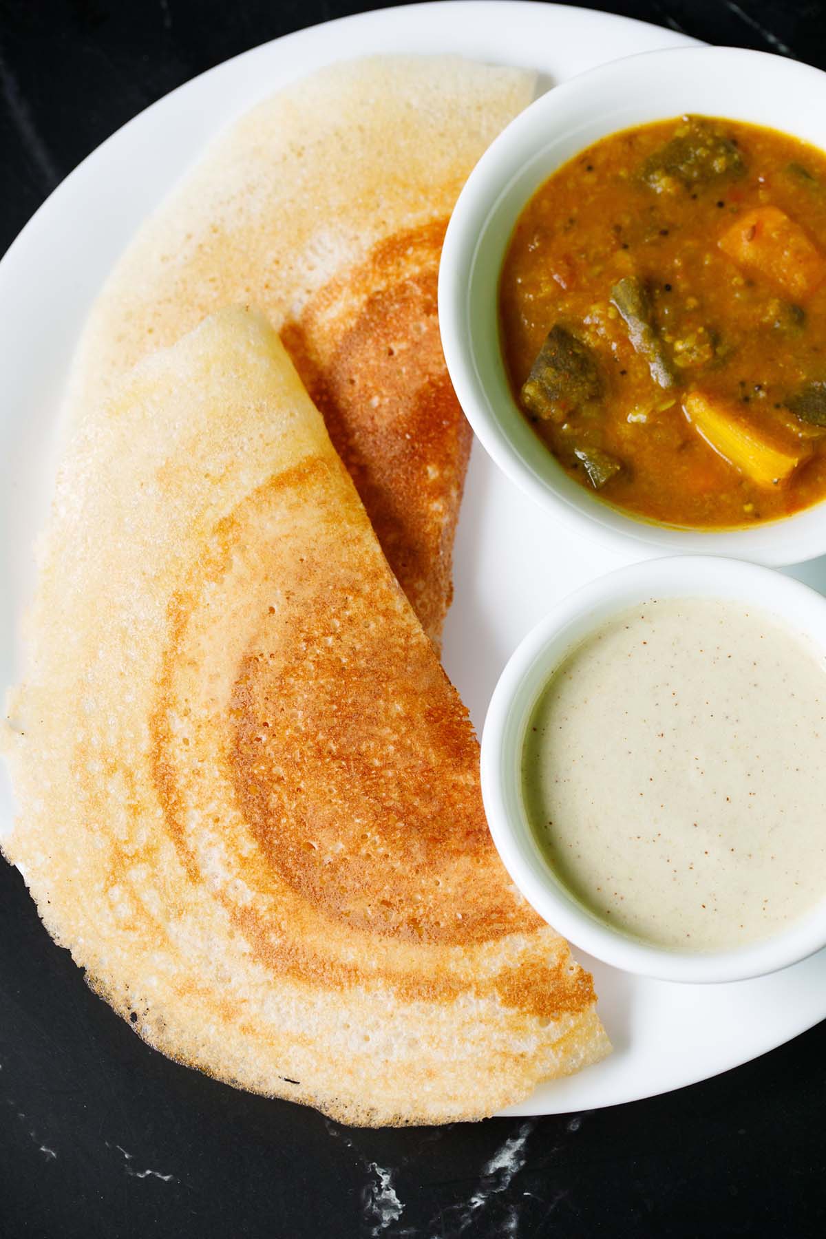 two folded, golden, crisp placed next to white bowls filled with sambar and coconut chutney on a white plate