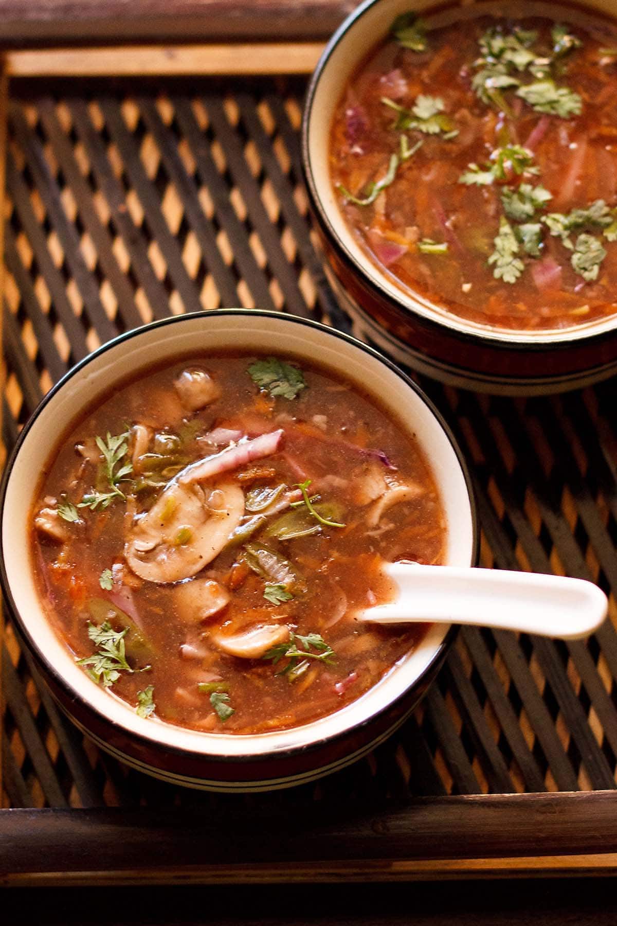 hot and sour soup with coriander garnish in a soup bowl with a white spoon inside on a dark brown bamboo tray