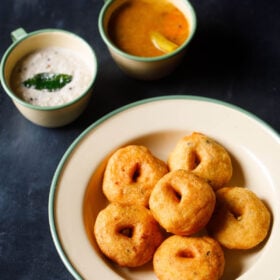 top shot of medu vada stacked neatly in green lined beige colored plate with cups filled with coconut chutney and sambar on the top