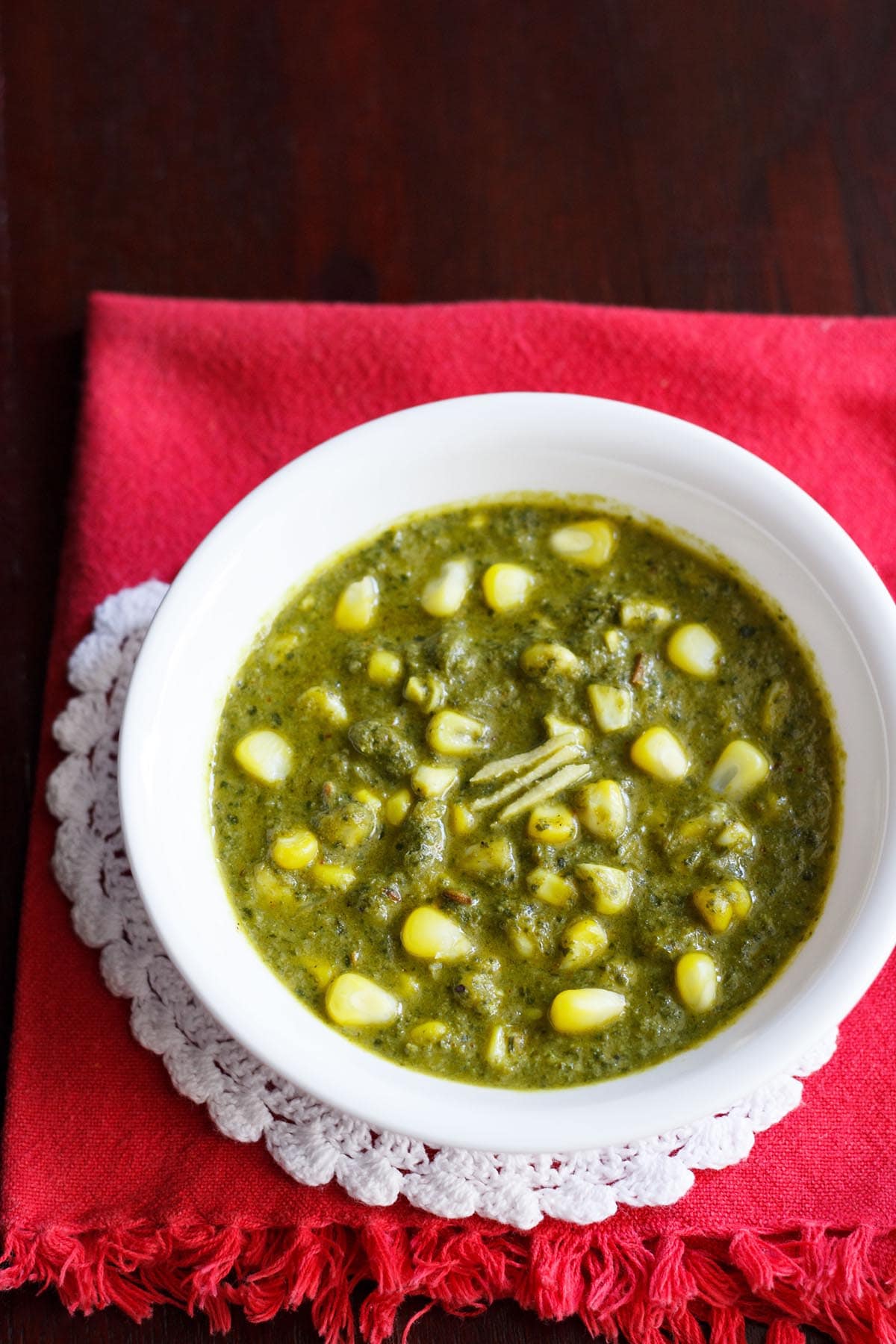 palak corn in a white bowl with three ginger juliennes on a white doily placed above folded red napkin