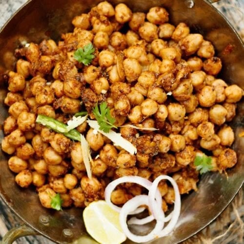 closeup shot of pindi chana in a metallic wok garnished with onion slices, cilantro, lemon wedge and green chilli strips