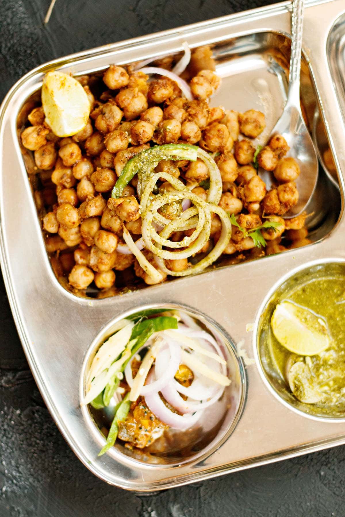 pindi chole served in a partitioned square tray with onion slices and lemon wedge on top with other partitions filled with onions, green chillies, mango pickle, lemon wedge and green chutney
