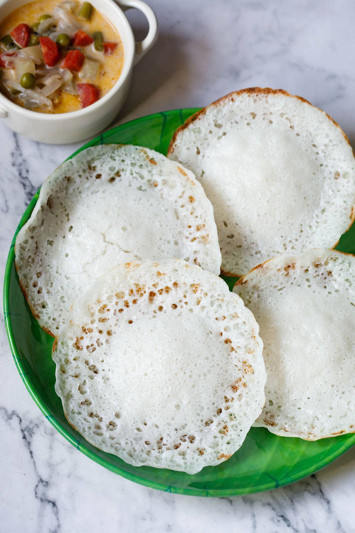 overhead shot of kerala appams on a green plate with vegetable stew in white bowl placed on top