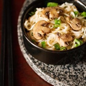 mushroom noodles with scallion greens in black bowl on a round grey black granite board