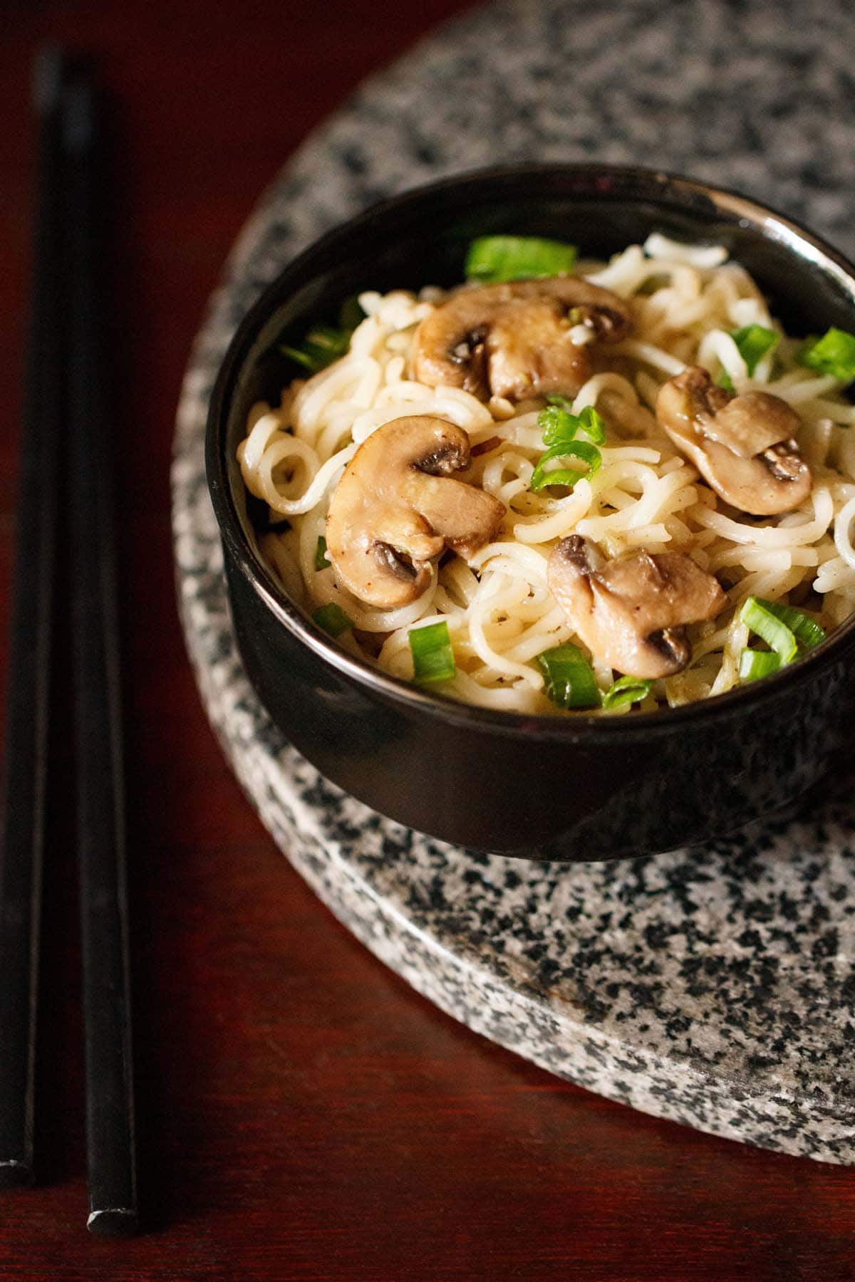 mushroom noodles with scallion greens in black bowl on a round grey black granite board