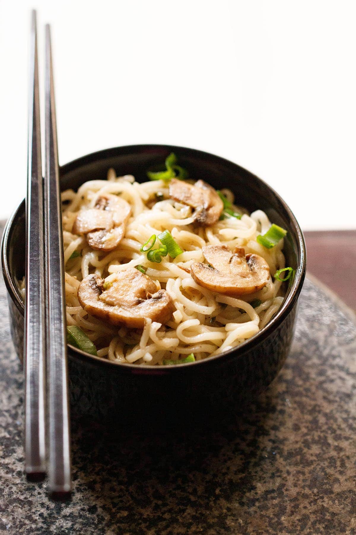 mushroom noodles with black chopsticks placed on top of black bowl on a round grey black granite board