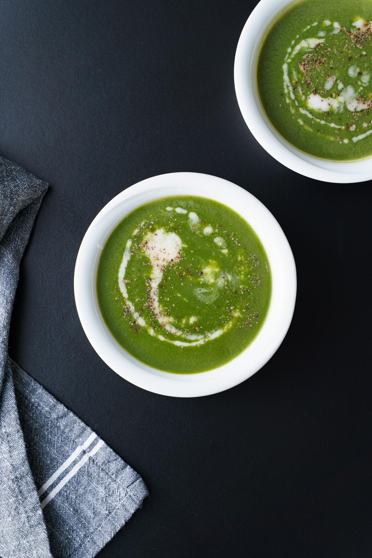 overhead shot of palak soup drizzled with coconut cream on top in a white bowl on a black board