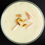 closeup overhead shot of phirni with pistachios and almond slivers garnish on glass bowl on black board
