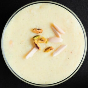closeup overhead shot of phirni with pistachios and almond slivers garnish on glass bowl on black board