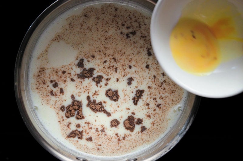 ground cardamom sprinkled above milk in pan with saffron infused milk being added from top