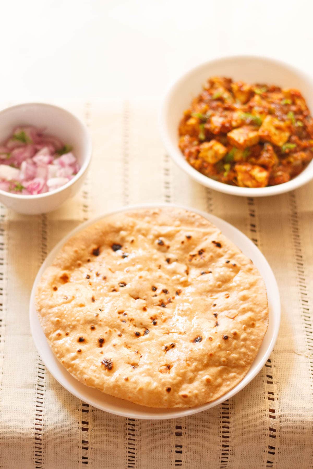 phulka smeared with ghee on a white plate with two white bowls of paneer curry and chopped red onions placed above on a cream cotton fabric