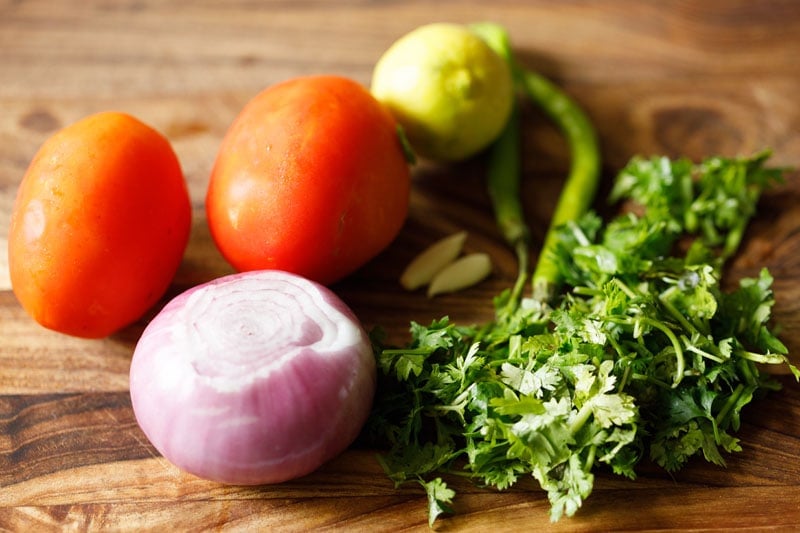 two tomatoes, peeled red onion, fresh cilantro, two cloves of garlic, two green chiles and a lemon on a cutting board