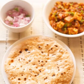 roti smeared with ghee on a white plate with two white bowls of paneer curry and chopped red onions placed above on a cream cotton fabric