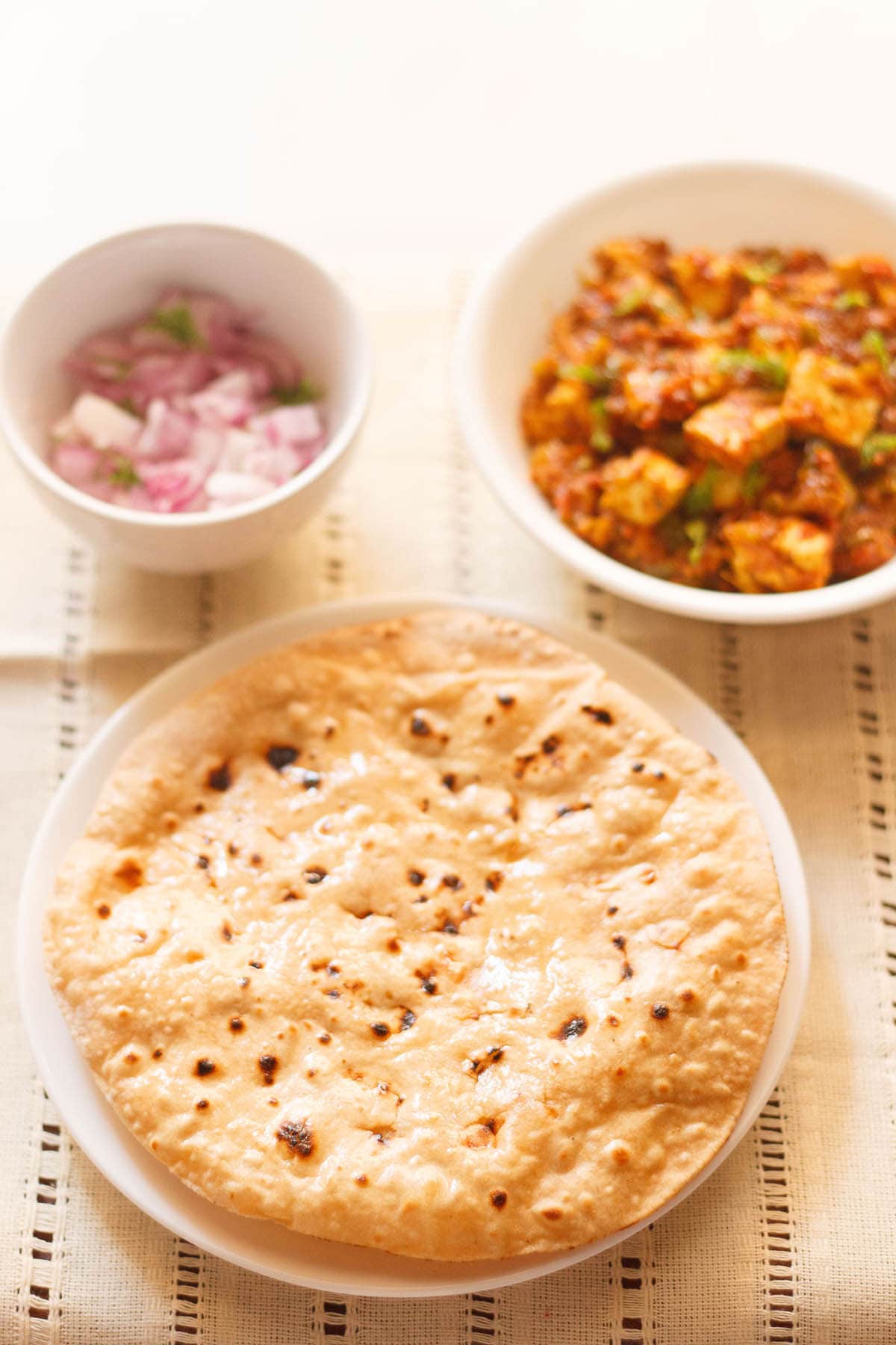 roti smeared with ghee on a white plate with two white bowls of paneer curry and chopped red onions placed above on a cream cotton fabric