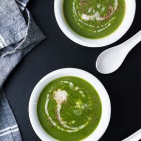 overhead shot of spinach soup topped with coconut cream in two white bowls and two white spoons placed on a black board