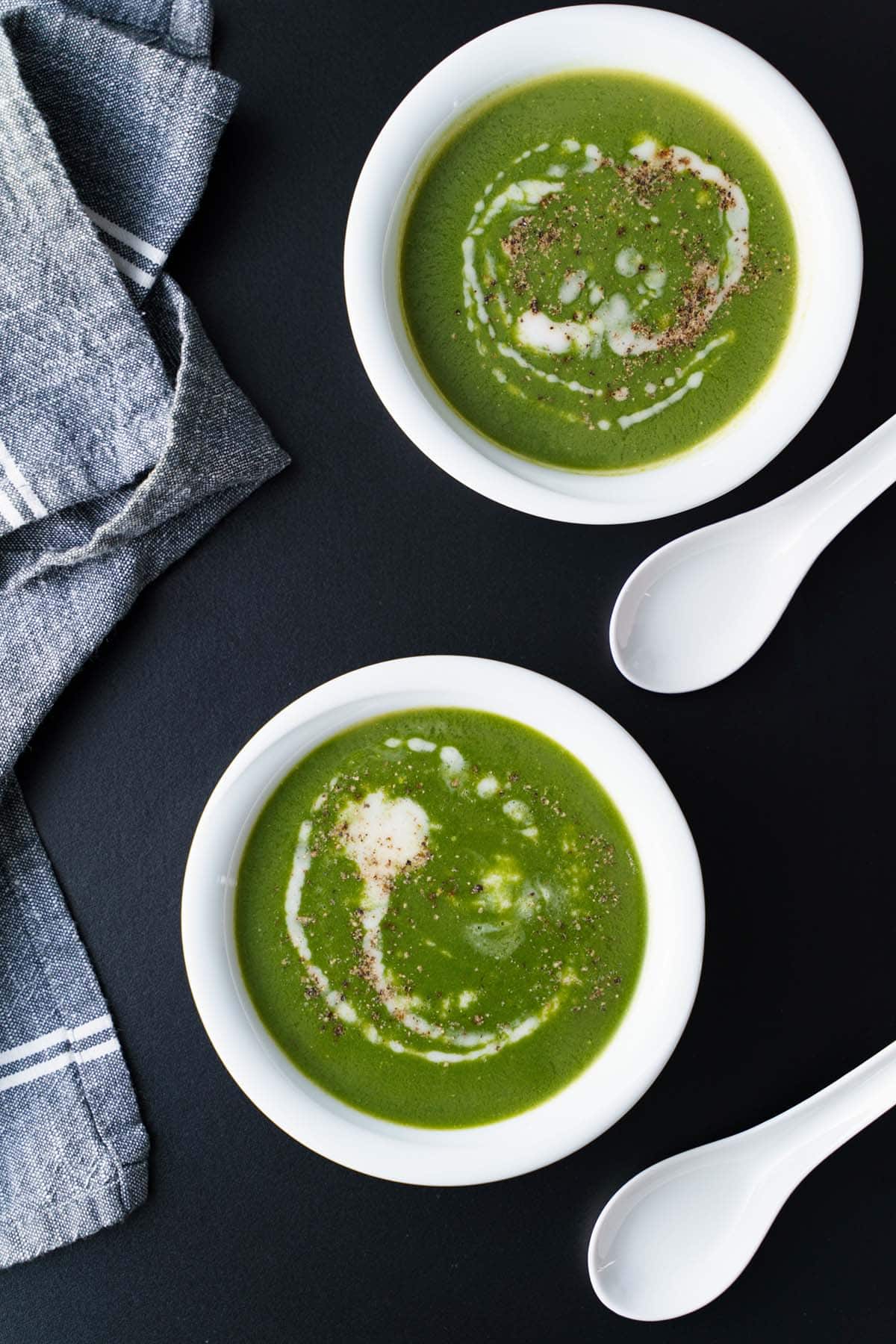 overhead shot of spinach soup topped with coconut cream in two white bowls and two white spoons placed on a black board