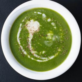 overhead shot of spinach soup drizzled with coconut cream and sprinkled with crushed black pepper in white bowl on a black board