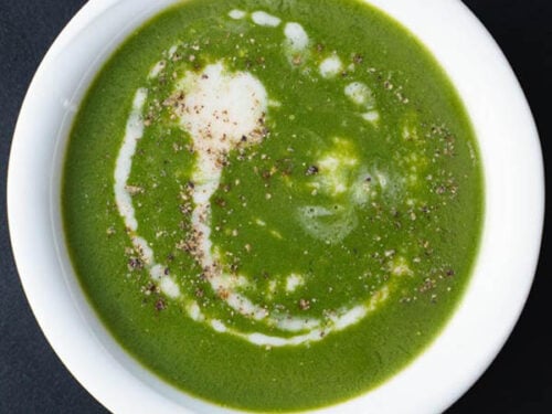 overhead shot of spinach soup drizzled with coconut cream and sprinkled with crushed black pepper in white bowl on a black board