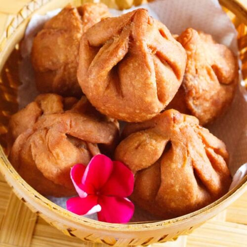 closeup shot of fried modak in a round bamboo bowl with a dark pink flower inside