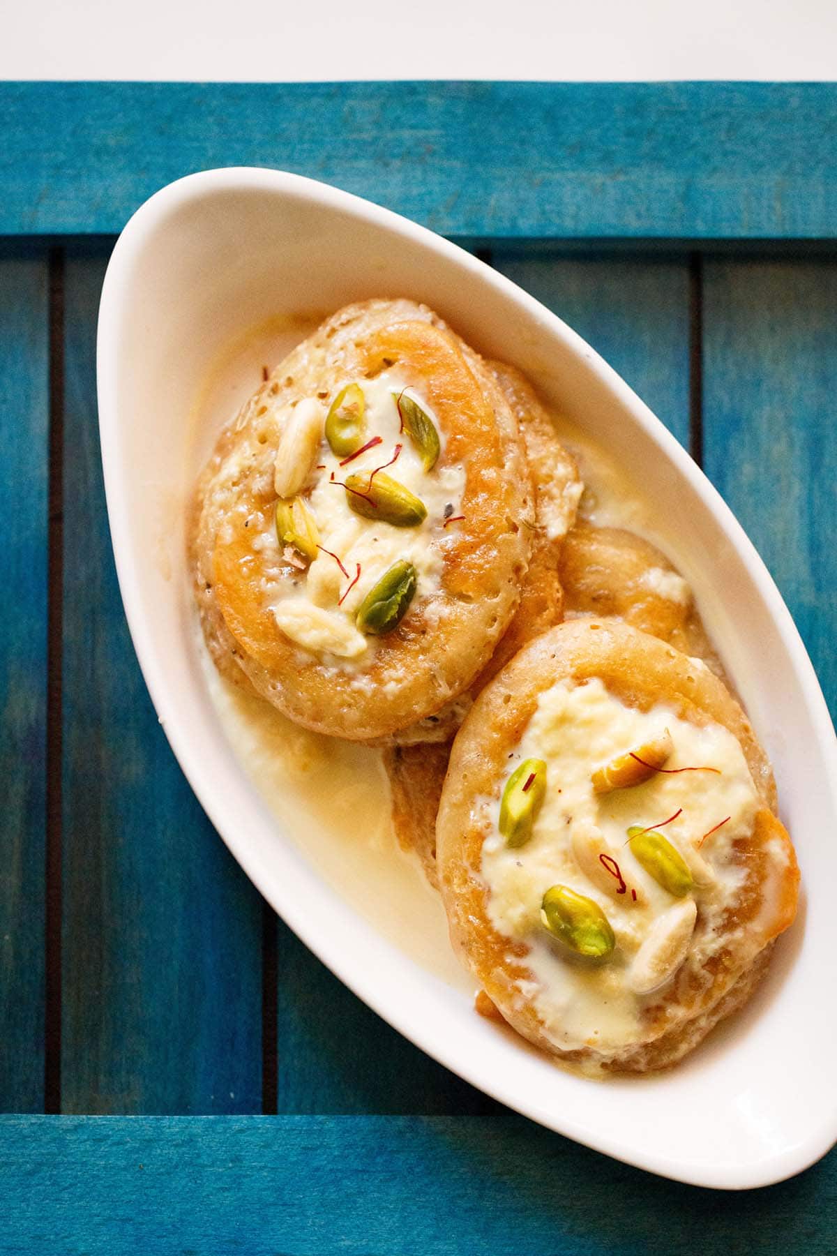 overhead shot of malpua with rabdi served in a oval plate on a dark blue tray