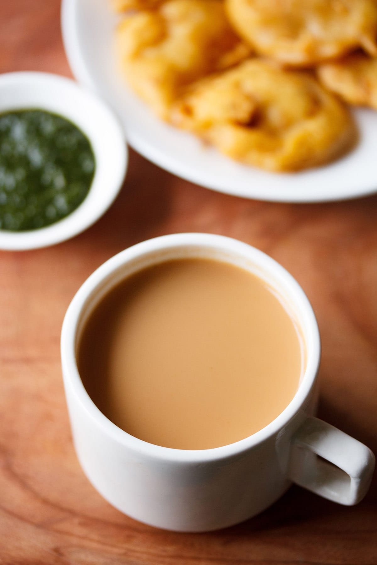 masala chai filled in a white cup with a side of onion pakora in white plate and green chutney in small white bowl