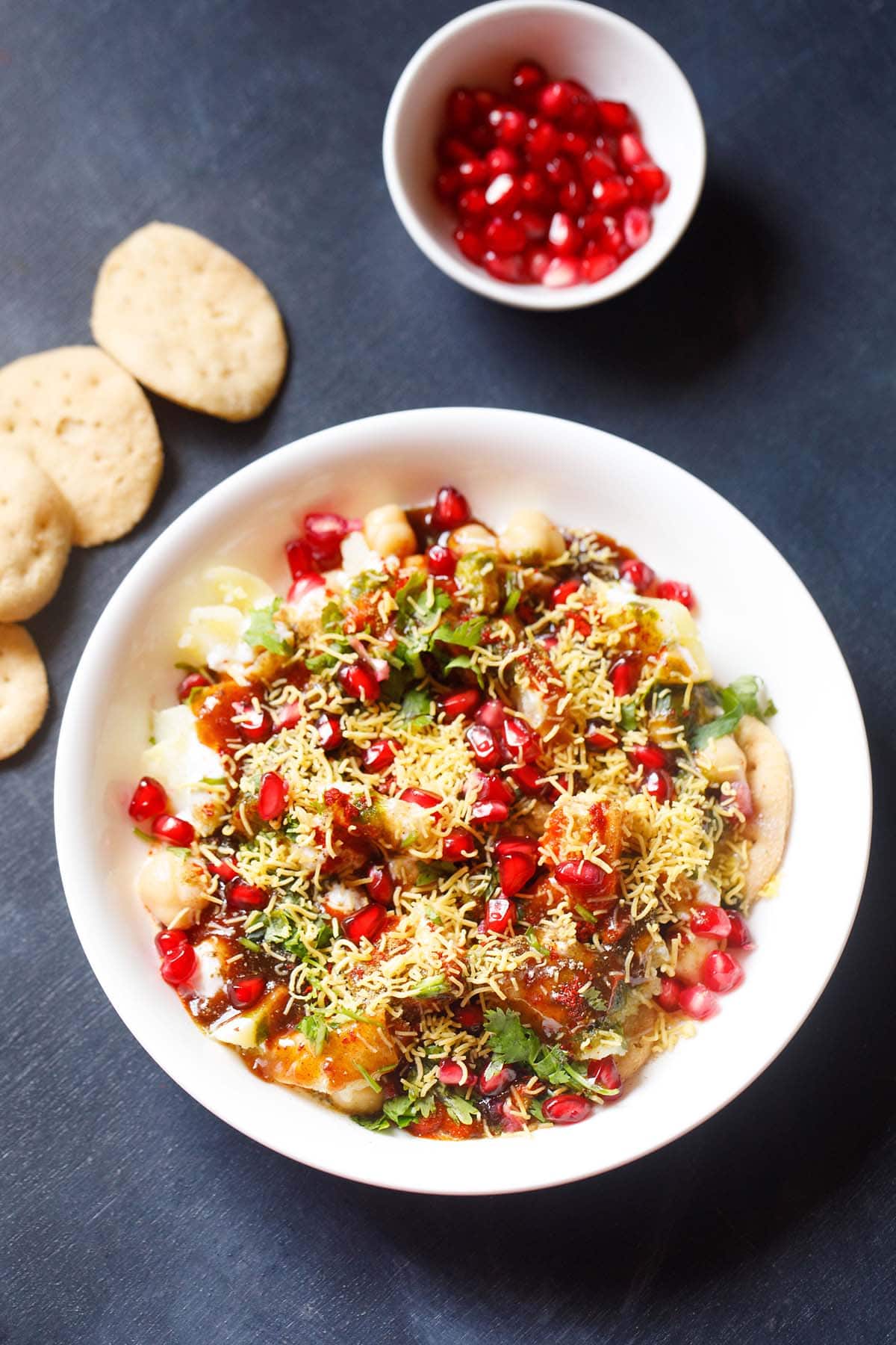 overhead shot of papdi chaat in a white shallow bowl with a side of papdi and pomegranate arils in a small white bowl