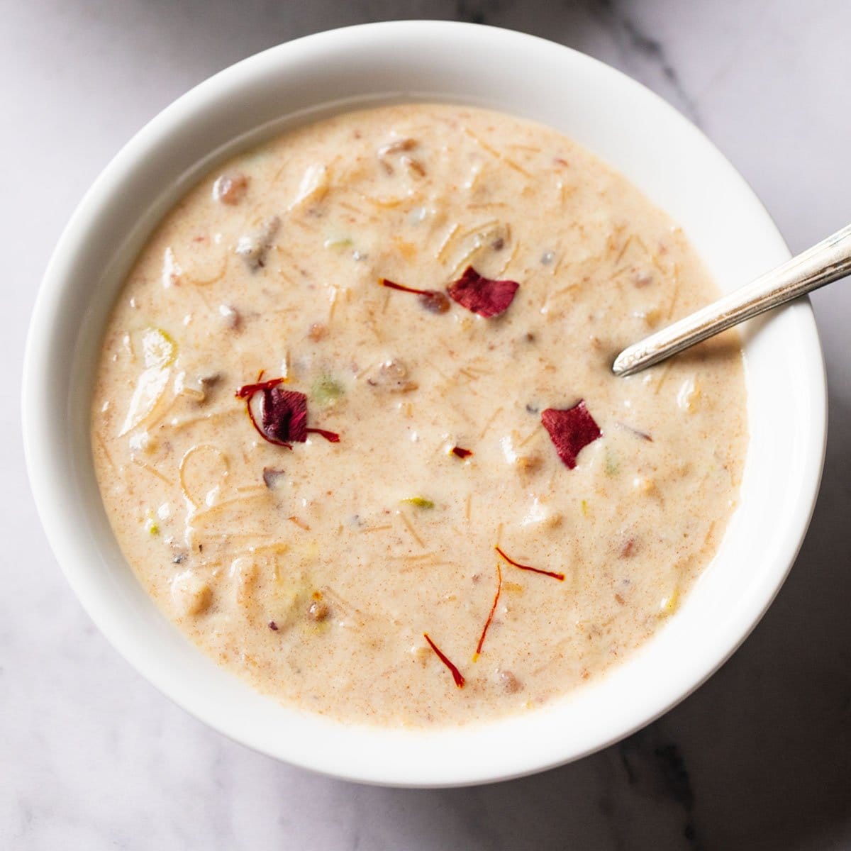sheer khurma garnished with rose petals in a white bowl with a brass spoon inside the bowl
