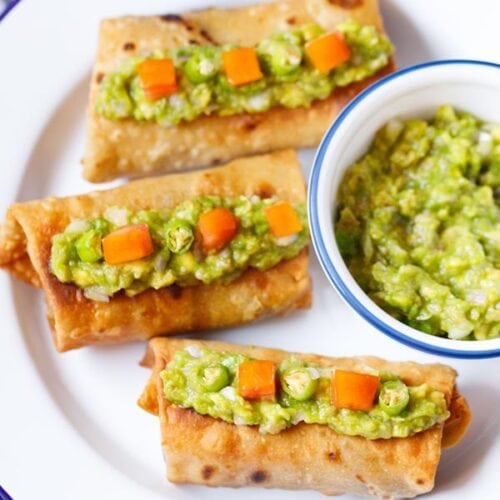 overhead shot of three chimichanga topped with guacamole and tomato cubes with a side bowl of guacamole on a white plate