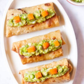 overhead shot of three chimichanga topped with guacamole and tomato cubes placed on an off-white oval tray