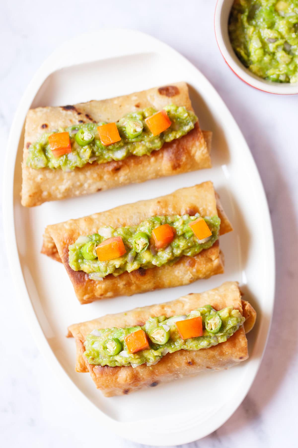 overhead shot of three chimichanga topped with guacamole and tomato cubes placed on an off-white oval tray