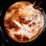 hot chocolate sprinkled with cocoa powder in a black mug with a dessert spoon inside