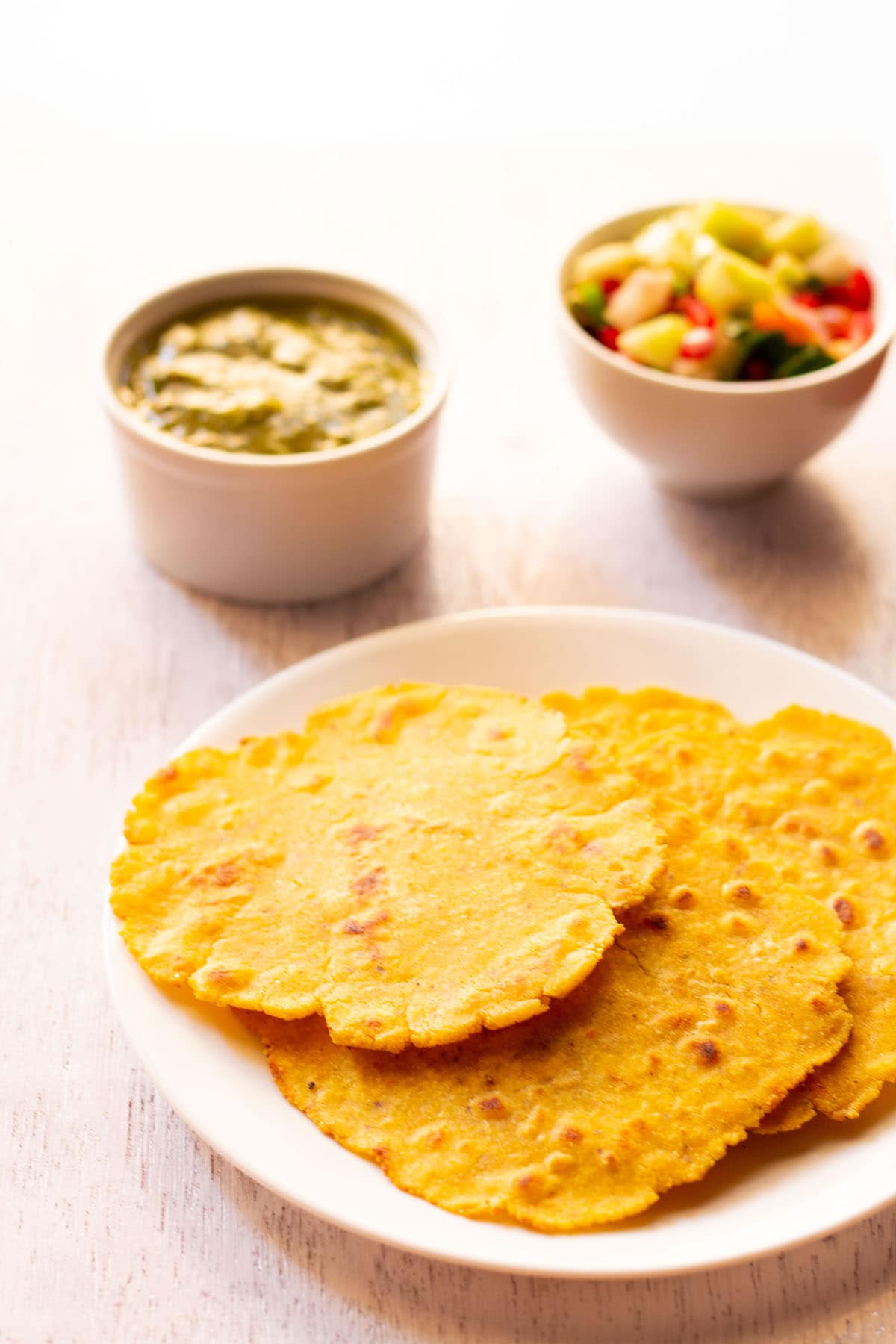 three makki di roti in a white plate with saag and a salad in white bowls placed on top side