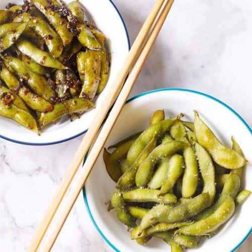 bowl of japanese salted edamame and spicy garlic edamame in two white bowls with chopsticks in center of bowls.