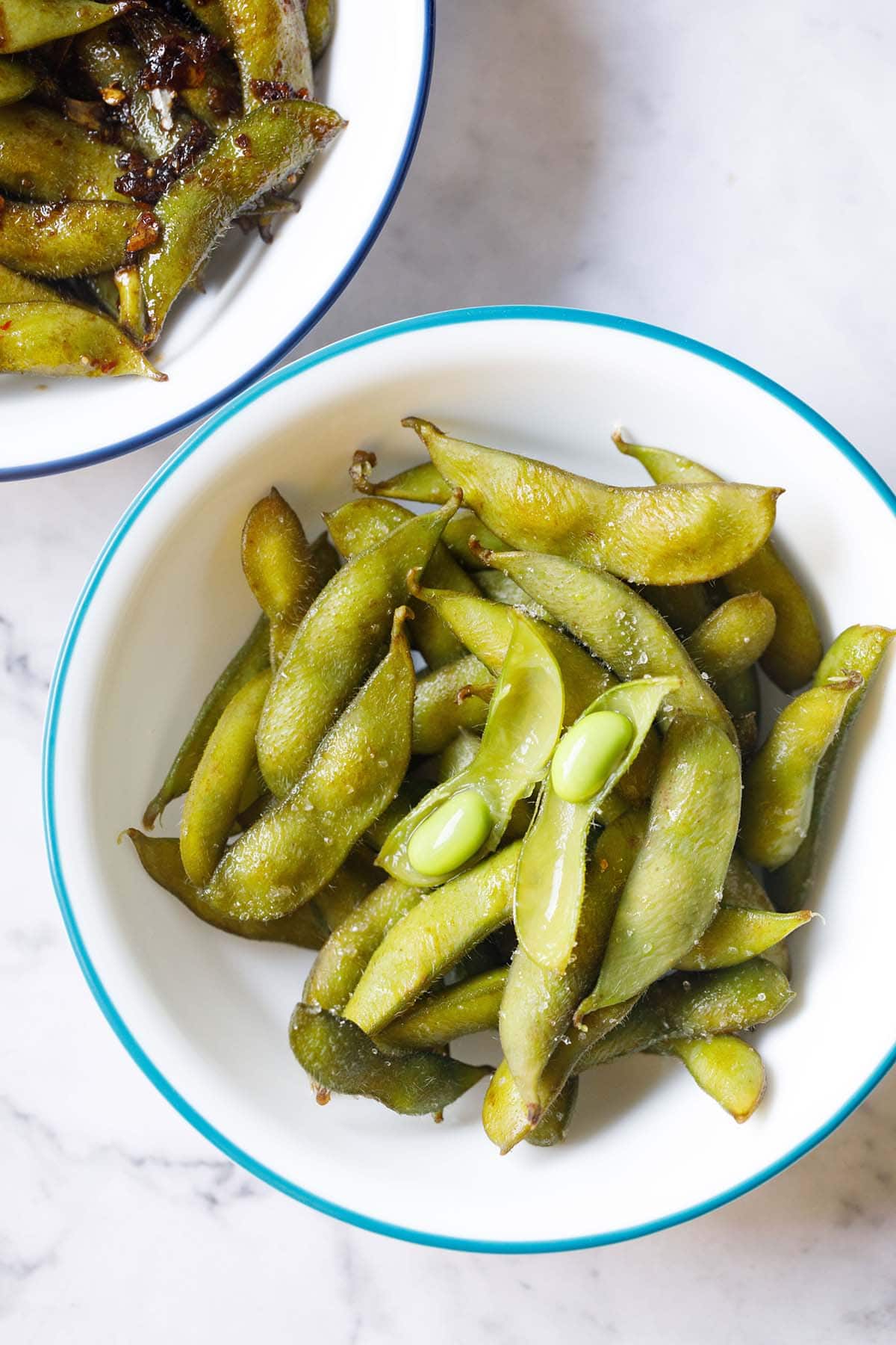 japanese style steamed and salted edamame beans with one edamame pod opened, in blue rimmed white bowl.