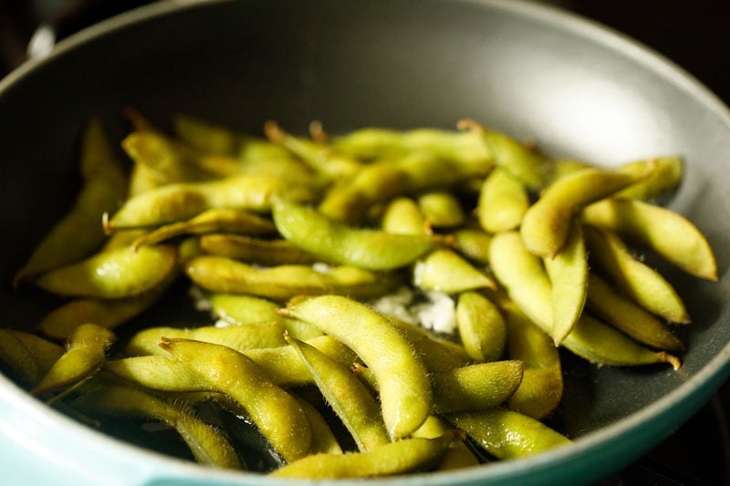 boiled edamame pods added to garlic in pan.
