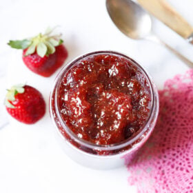 overhead shot of easy homemade strawberry jam in a clear glass jar with two strawberries and a pink crocheted doily on the table.