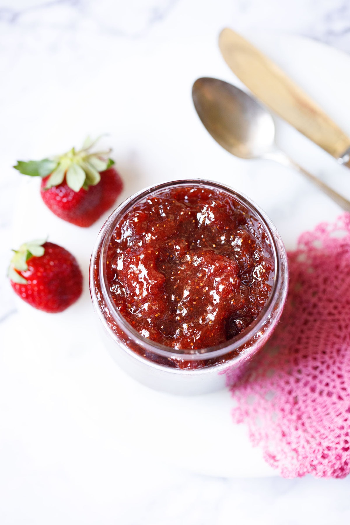 overhead shot of easy homemade strawberry jam in a clear glass jar with two strawberries and a pink crocheted doily on the table.
