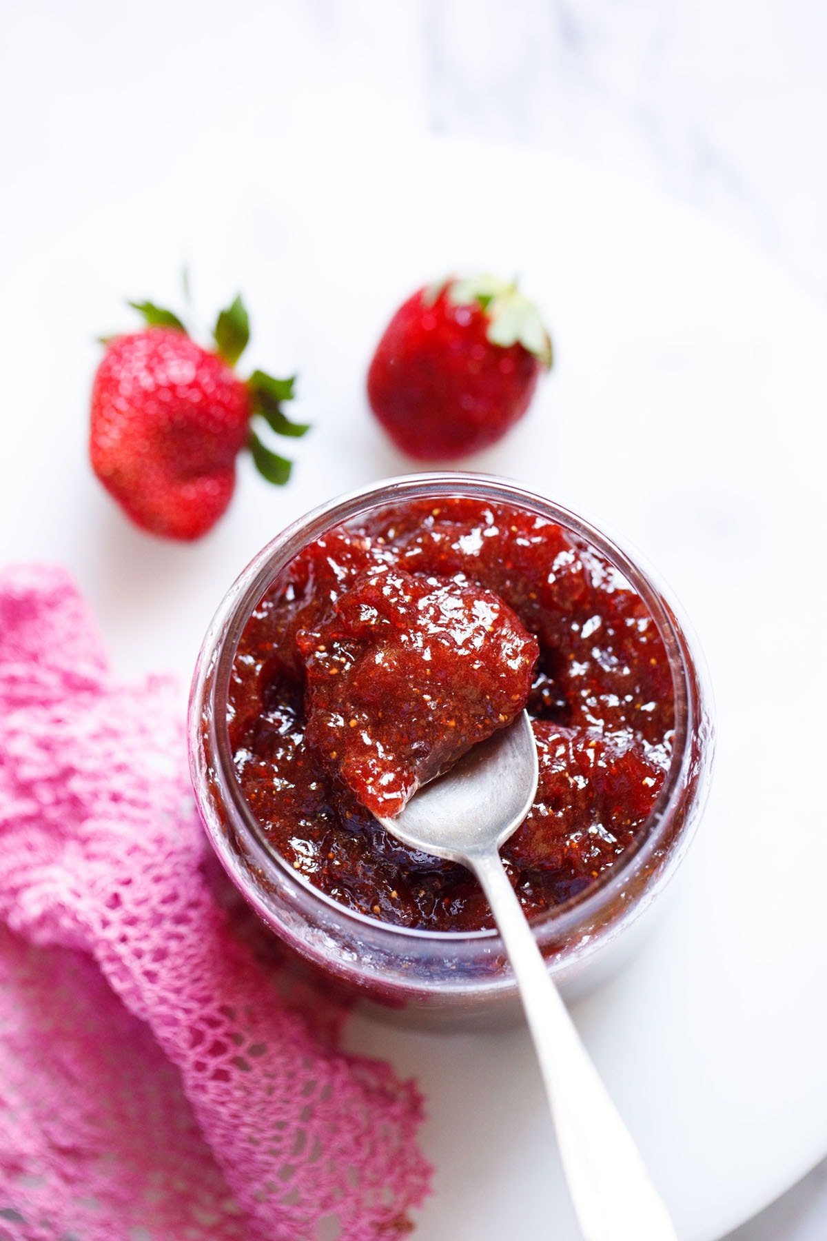 overhead shot of glass jar of strawberry jam on a white table with a silver spoon showing the consistency of it.