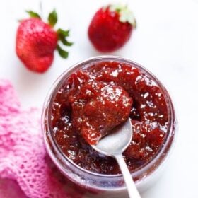 overhead shot of glass jar of strawberry jam on a white table with a silver spoon showing the consistency of it with text layovers.