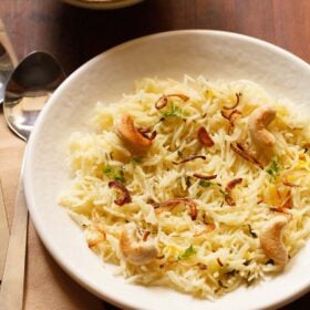 biryani rice garnished with fried cashews and caramelized onions on an off white plate with a spoon and paneer curry in a bowl