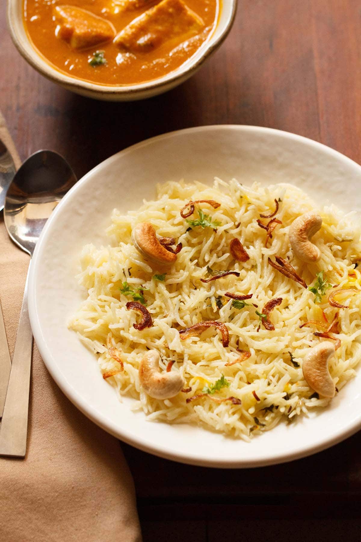 biryani rice garnished with fried cashews and caramelized onions on an off white plate with a spoon and paneer curry in a bowl