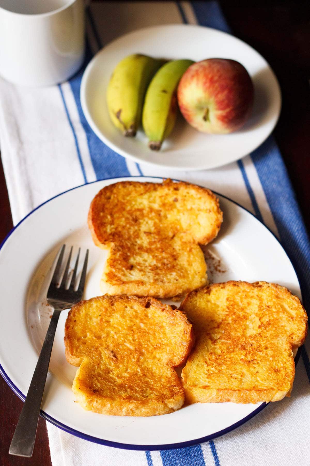 three french toast served on a plate with two bananas and apple on a second white plate.