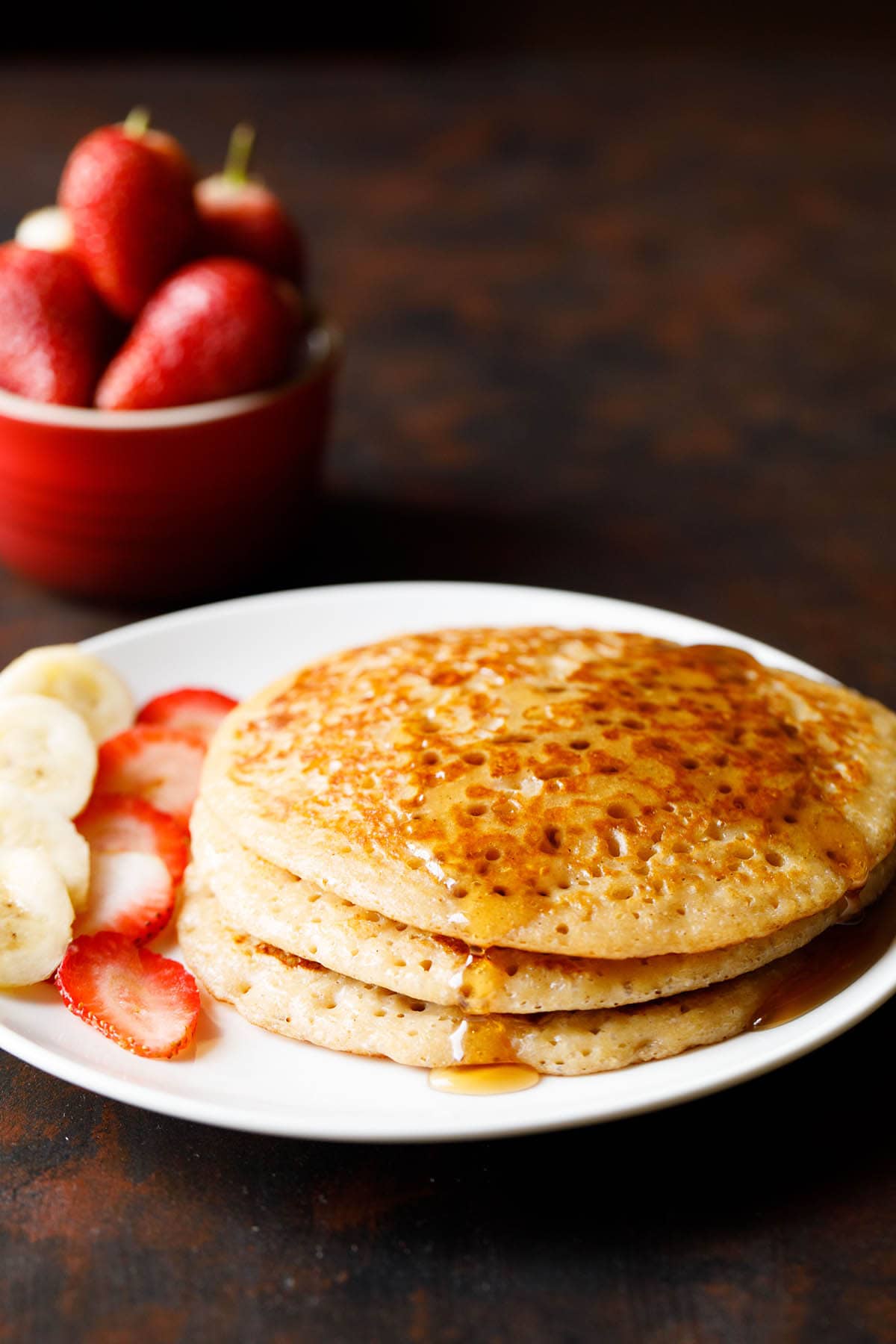 stack of pancakes with maple syrup drizzled and sliced bananas and strawberries on white plate.