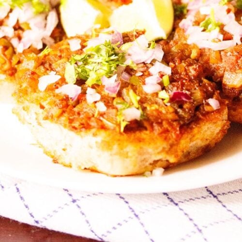 masala pav garnished with chopped onions, coriander leaves and served on a white plate with lemon wedges on top.