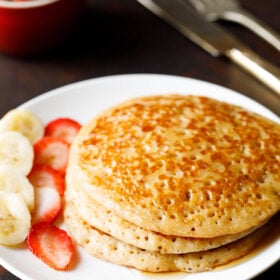 stack of pancakes with maple syrup drizzled and sliced bananas and strawberries on white plate.