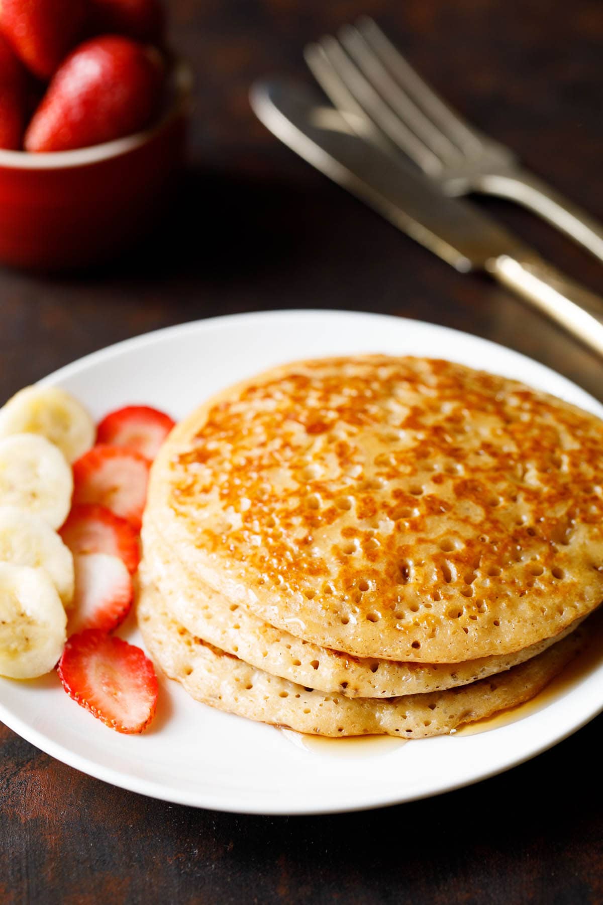 stack of pancakes with maple syrup drizzled and sliced bananas and strawberries on white plate.