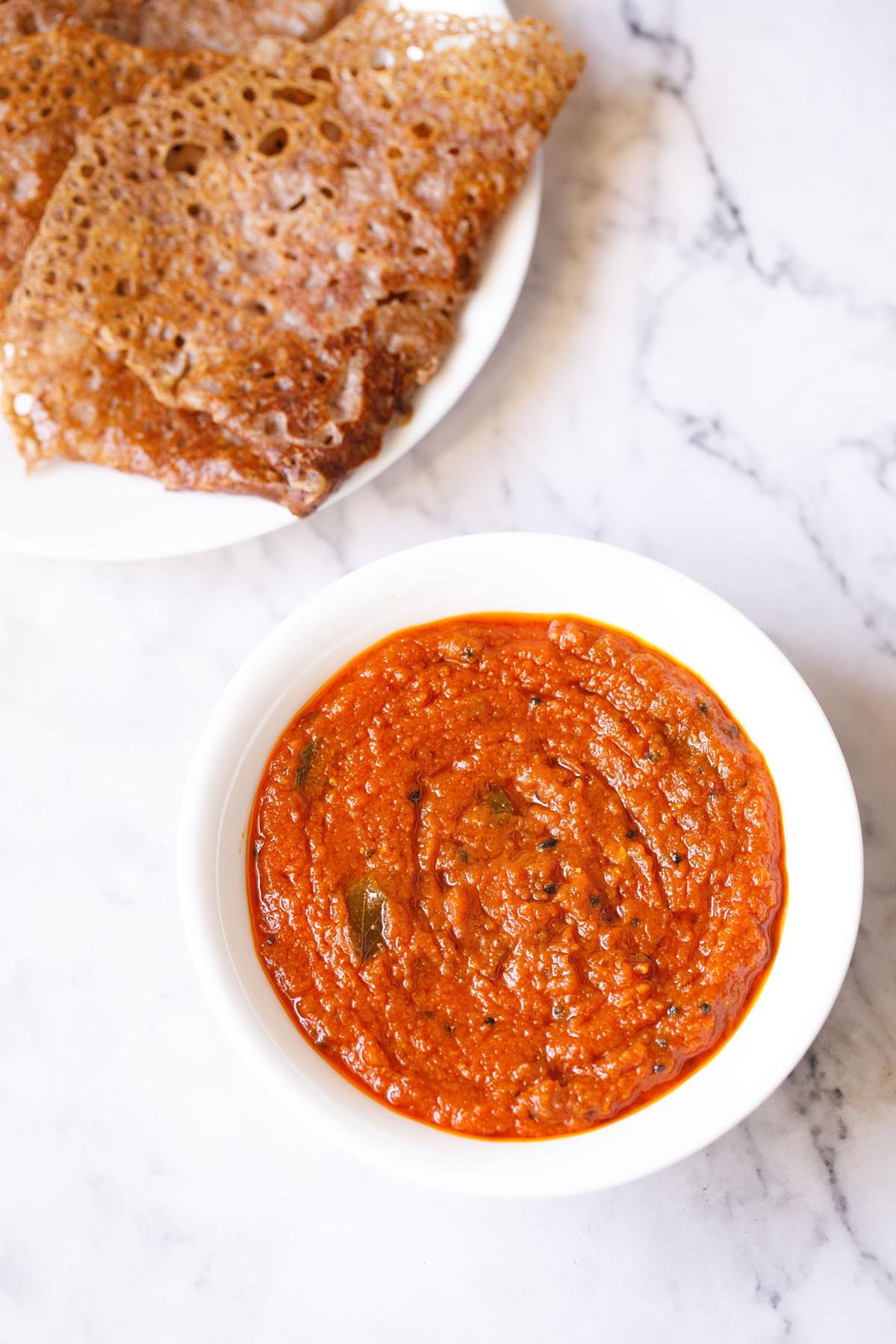 tomato pickle or tomato thokku in white bowl with ragi dosa in a white plate above.