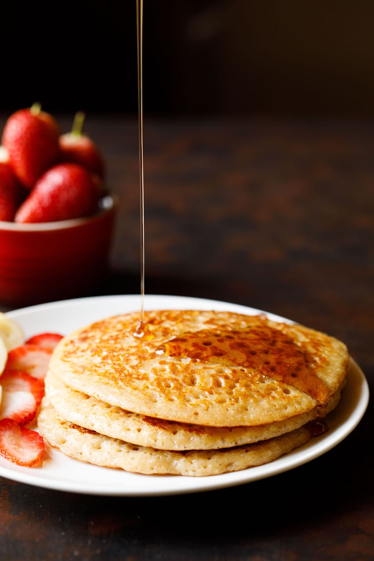 whole wheat pancakes stack in a plate with maple syrup being poured on top.
